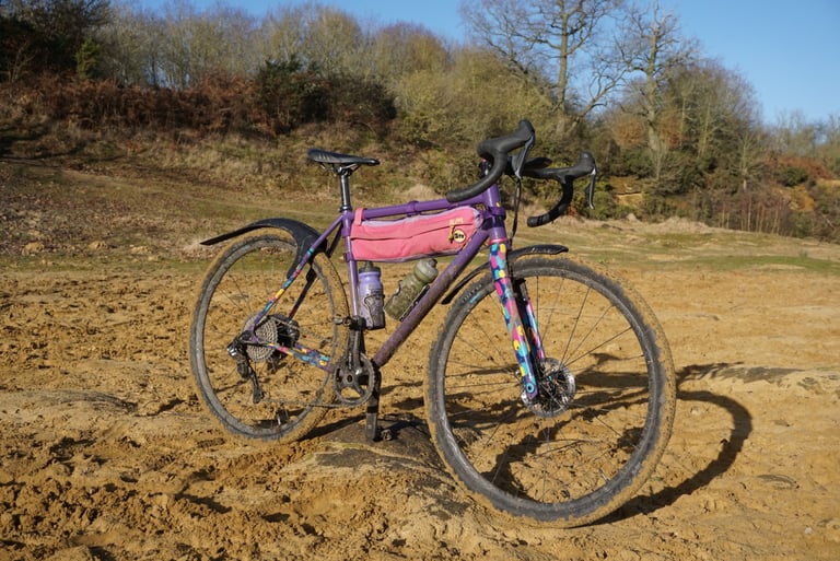Gravelhugger mudguards mounted on a muddy purple gravel bike with a pink frame bag, parked on a sandy track.
