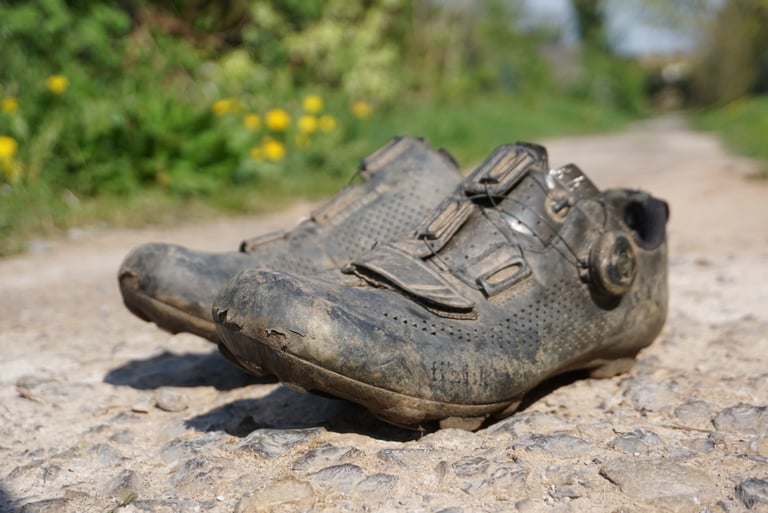Well-used Fizik Terra X5 shoes, scuffed and muddy, on a rough gravel track after two years of riding.