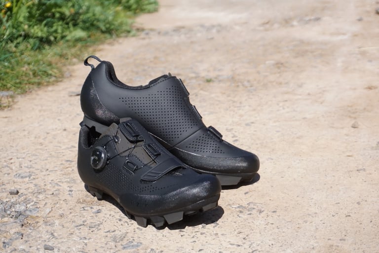 Black Fizik Terra X5 shoes stacked on a dirt track, showing the perforated upper and BOA dial.