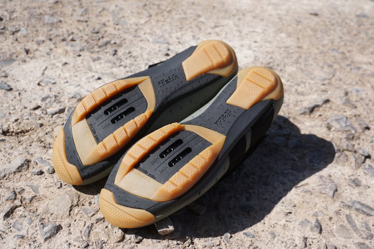 Soles of the Fizik Terra Powerstrap X4 with gum rubber tread either side of the SPD cleat slot, on a stony track.