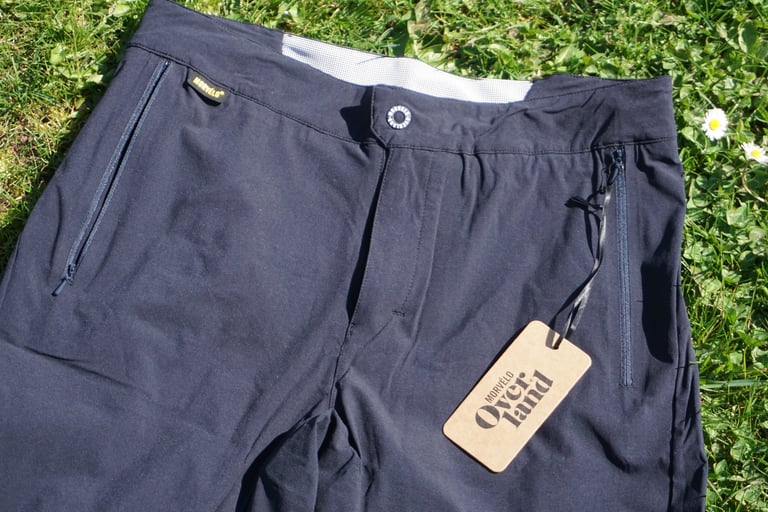 Overland Selector shorts flat on grass, showing the zipped side pockets, front popper and hang tag.