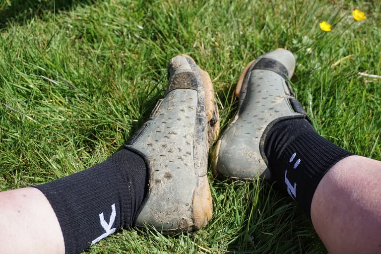 Muddy Fizik Terra Powerstrap X4 shoes on a rider's feet, resting on grass after a ride.