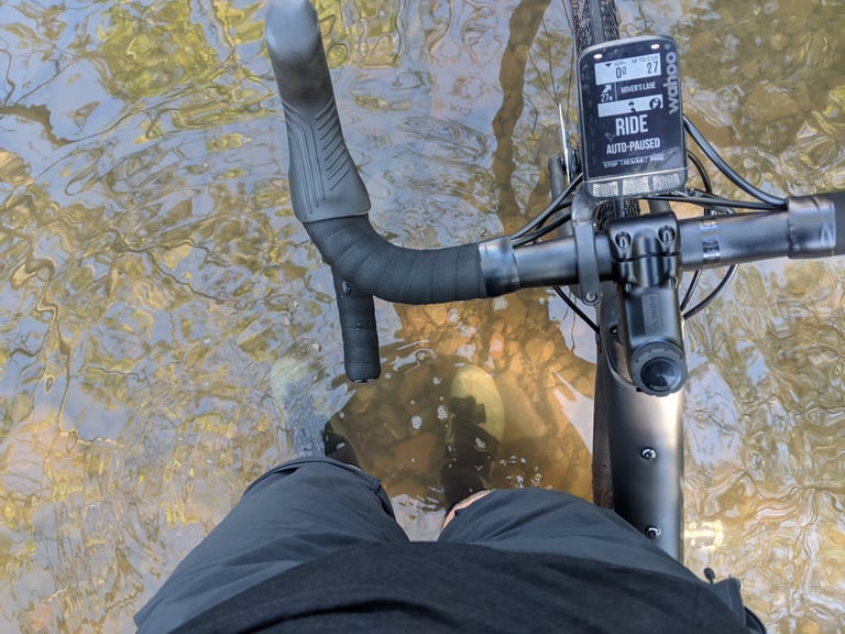 Rider's view down from a gravel bike standing in a shallow stream, Fizik Terra Powerstrap X4 shoes under the water.