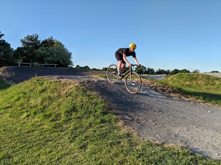 Rider in a yellow helmet and Overland Selector shorts rolls a gravel bike over a pump-track mound under a blue sky.