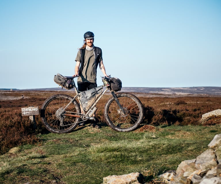 Matty Waudby stands with a muddy bikepacking rig on heather moorland beside a Goathland signpost.