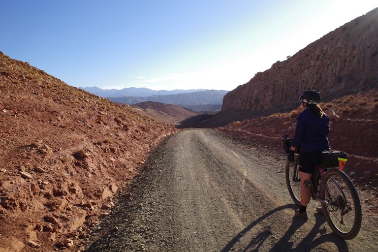 Shona from Keep Pedalling stands over a loaded touring bike on a gravel road between red rocky hills in Morocco.