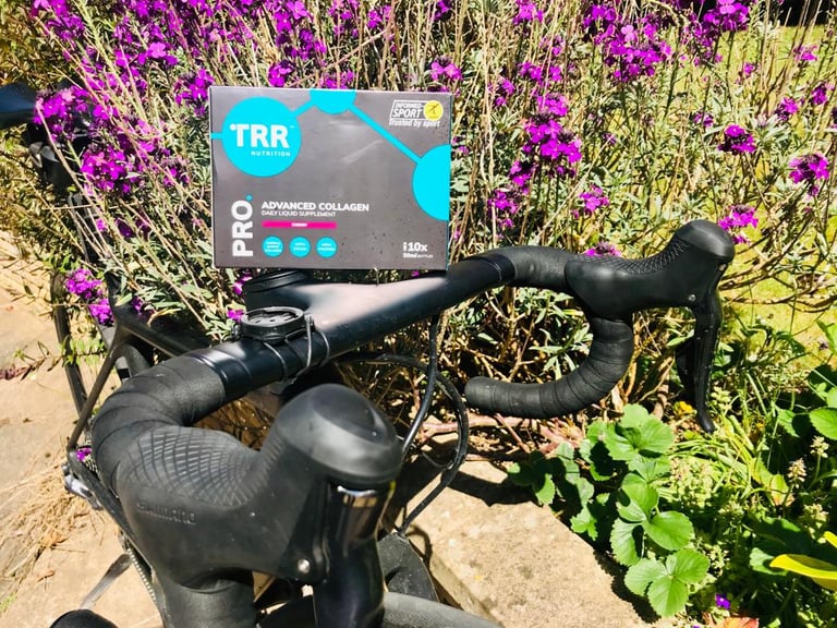 Box of TRR Nutrition Pro Advanced Collagen resting on the handlebars of a drop-bar bike beside purple flowers.