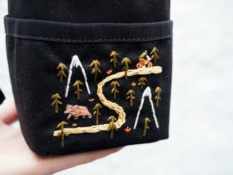 Close-up of the hand-embroidered bear, trail and mountains on the Get Wild Voila! snack bag in waxed canvas.