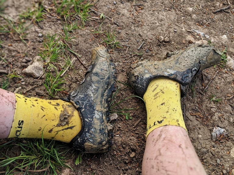 Mud-caked Specialized Recon shoes and yellow socks on a dirt track after a hike-a-bike section.
