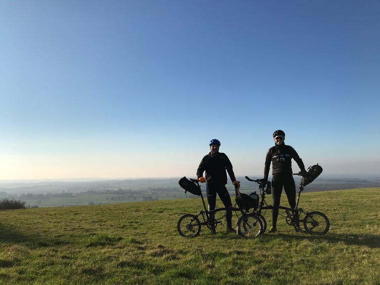 Mark and Jack stand with their Brompton folding bikes on a grassy hilltop under a clear sky before the TransAtlantic Way.