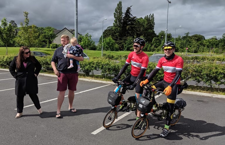Two riders in red Brompton jerseys on loaded Bromptons at the start of the TransAtlantic Way, family watching in a car park.