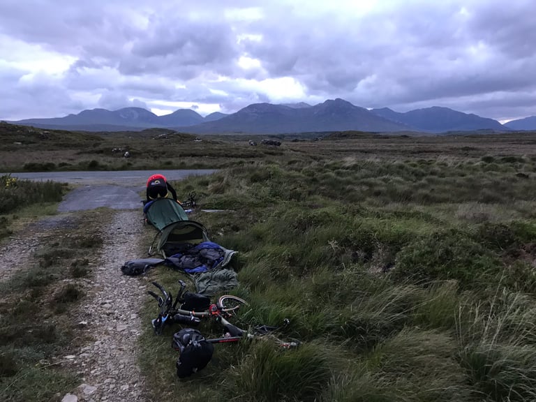 Two bivvy bags and loaded Bromptons strewn beside a bog road at dawn, the Twelve Bens beyond, on the TransAtlantic Way.