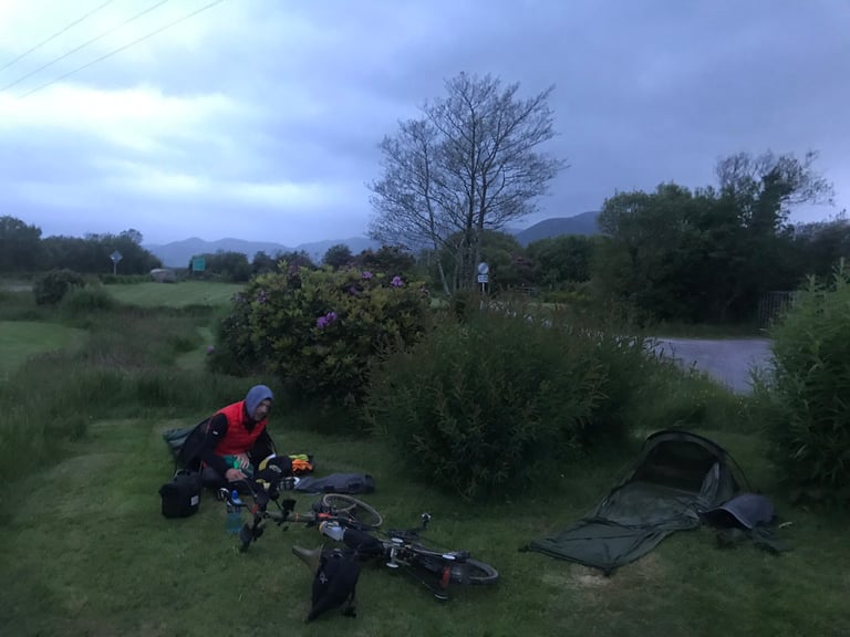 Rider sorts kit beside a hooped bivvy and a Brompton lying on wet grass at a damp TransAtlantic Way camp.