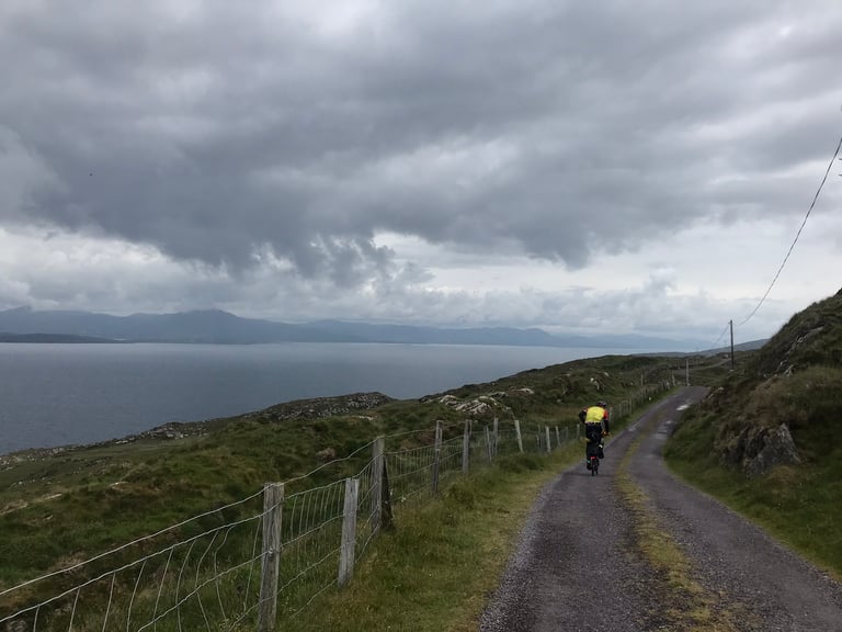 Brompton rider heads up a narrow lane above a wide bay under heavy cloud on the last day of the TransAtlantic Way.