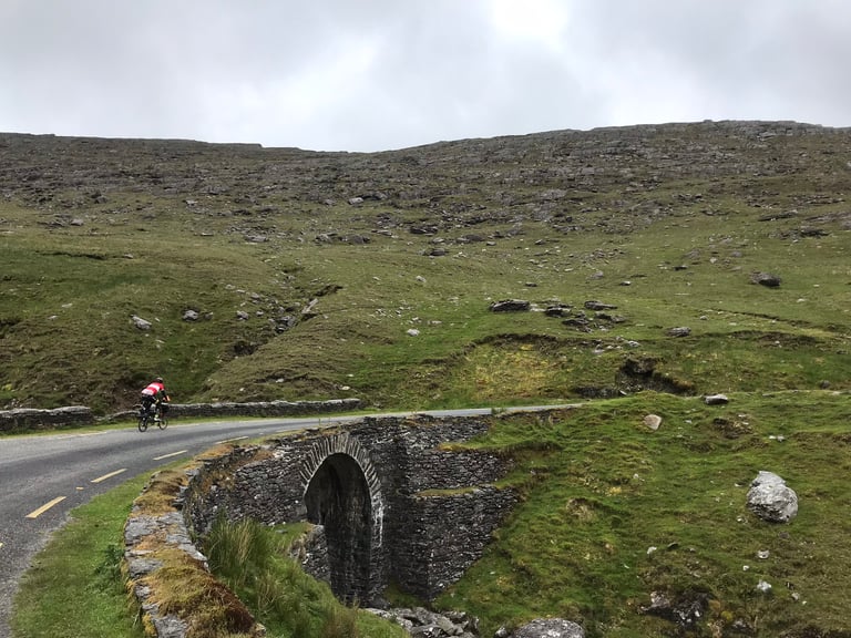 TransAtlantic Way rider on a Brompton rides a quiet road past a stone arch bridge on the Beara Peninsula, Ireland.