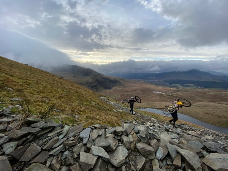 Two riders carry bikes over their shoulders on a rocky path up Snowdon, with a lake and cloud-hung hills below.