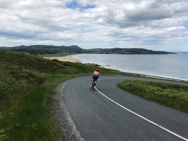 Brompton rider sweeps round a downhill bend above a sandy beach and bay on the TransAtlantic Way in Ireland.