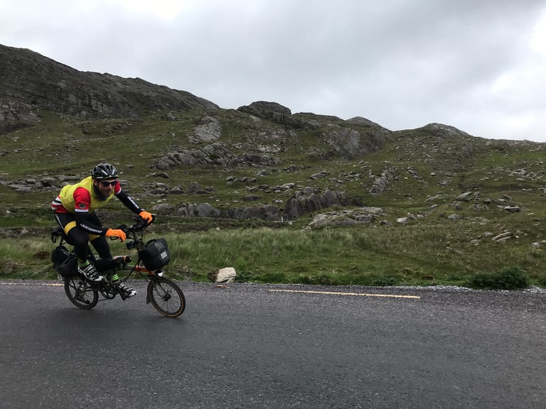 TransAtlantic Way rider on a loaded Brompton with aerobars rides fast past rocky hillside in Ireland.