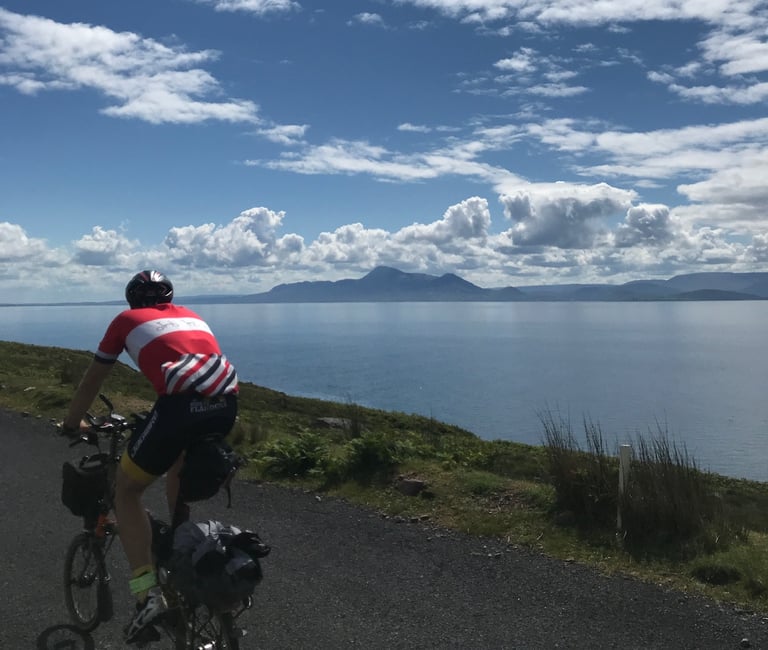 Brompton rider with loaded bags cycles beside a calm bay under blue sky and cloud on the TransAtlantic Way.