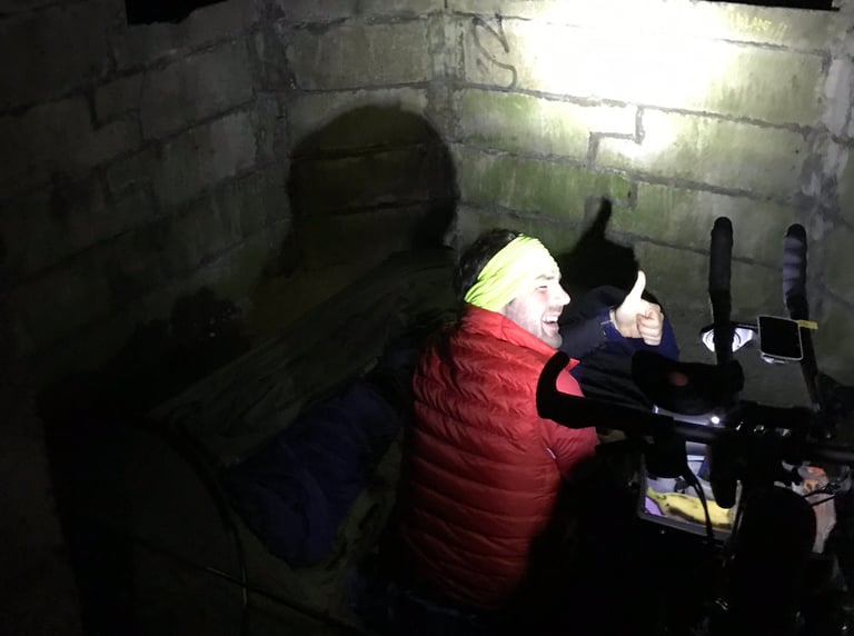 Rider in a red gilet and yellow headband gives a thumbs up in a concrete hut used as a bivvy near Malin Head.