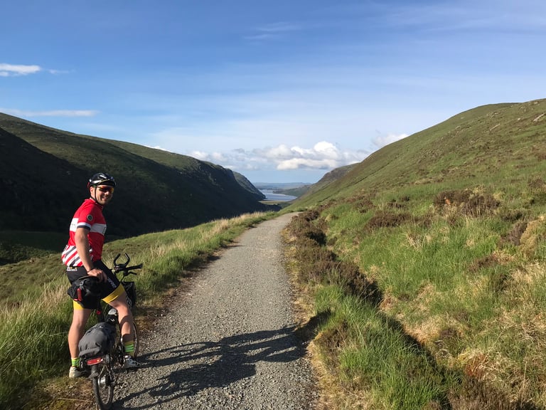 Rider on a Brompton pauses on a gravel track above a valley and lake, before the descent into Glenveagh National Park.