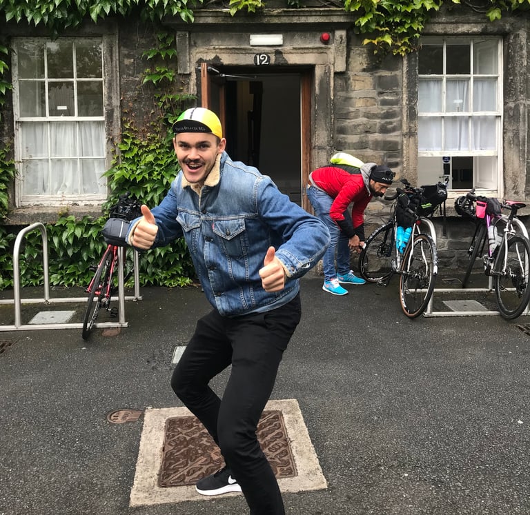 Rider in a TransAtlantic Way cap and denim jacket gives two thumbs up at registration in Dublin, bikes parked behind.
