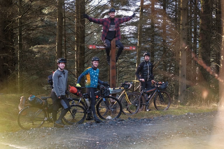 Four bikepackers with loaded bikes pose at a forest waymarker, one perched on top of the post.