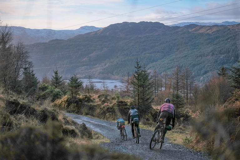 Three bikepackers ride a loose gravel descent towards Loch Long, with forested hills beyond.