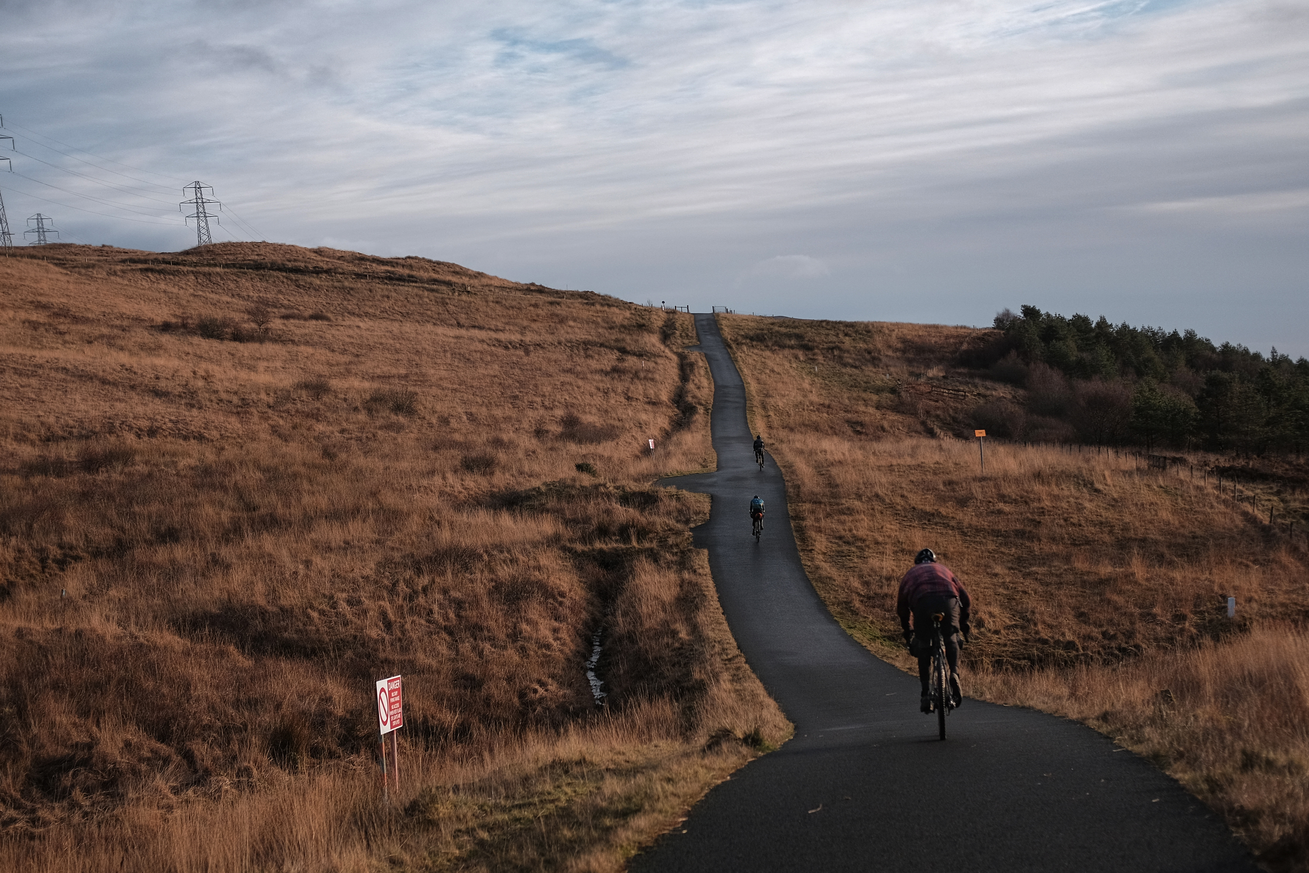 Three riders climb a narrow tarmac lane across open moorland, linking gravel sections of the Argyll route.