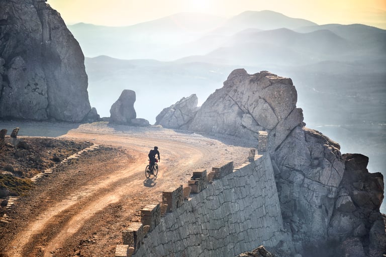 Lone gravel rider on a winding dirt road along a stone-walled cliff edge, with hazy mountains behind.