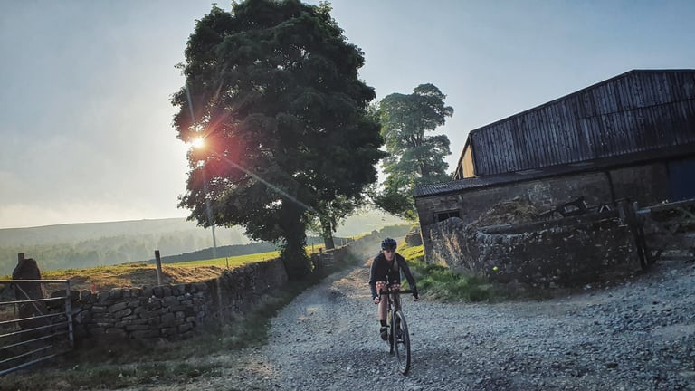 Early-morning sun flares through a tree as a rider passes a stone barn on a gravel lane in the Peak District.
