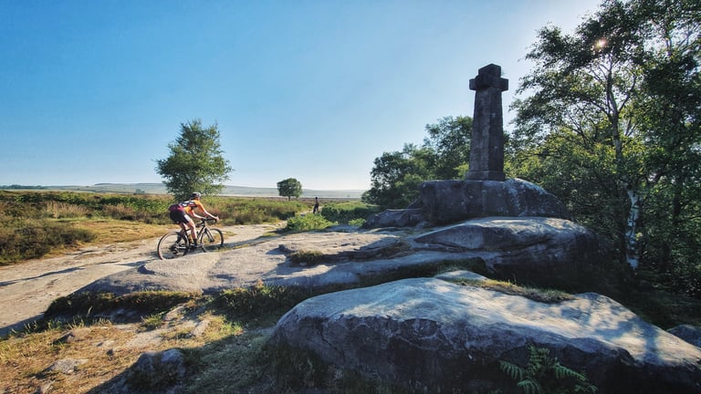 Dirty Kanzelled rider on a rocky Peak District moorland track passing a tall stone cross monument at sunrise.