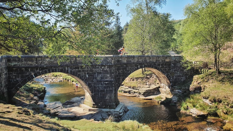 Rider crosses an old stone arch bridge over a shallow river in the Peak District on the Dirty Kanzelled route.