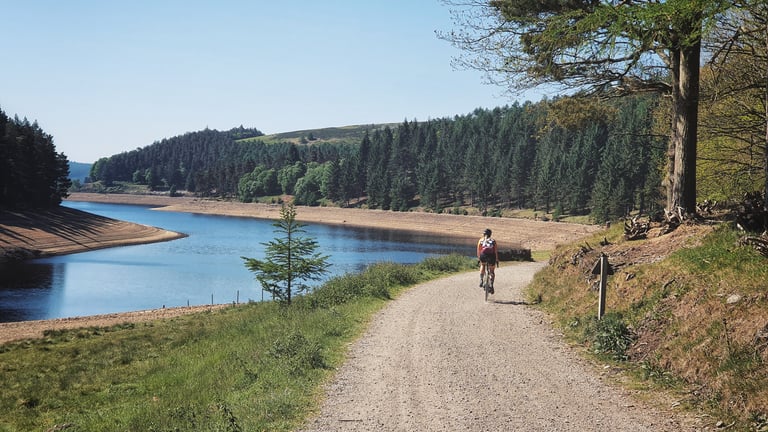 Rider on a gravel track beside Ladybower reservoir, with conifer woods on the far shore, during the Peak District ride.