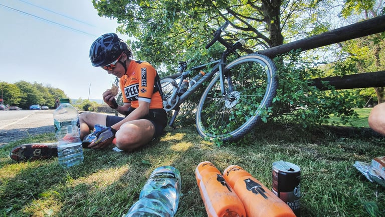 Magspeed rider eats a snack on the grass at a roadside refuel stop, gravel bike propped against a fence.
