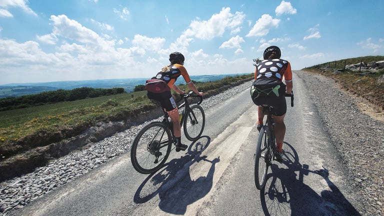 Two Magspeed Racing riders on a quiet lane with gravel verges above rolling Peak District hills on the Dirty Kanzelled.