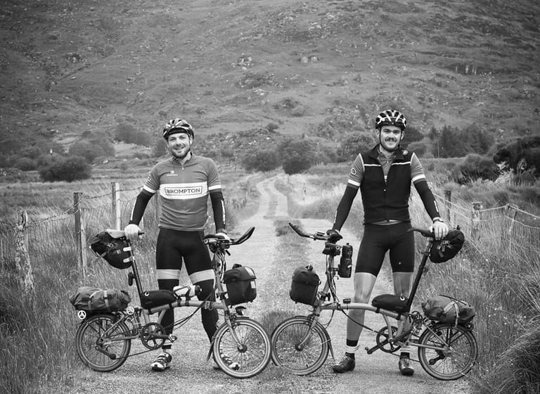 Mark and Jack stand beside their loaded Brompton folding bikes on a gravel track in Ireland, in black and white.