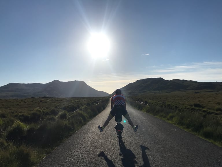 Brompton rider holds both legs out wide on a quiet road below mountains in Connemara, TransAtlantic Way Race.