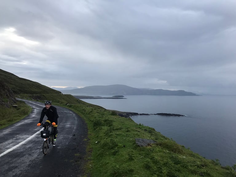 Smiling rider on a Brompton rounds a wet coastal road on Achill Island, grey sea and headlands beyond.