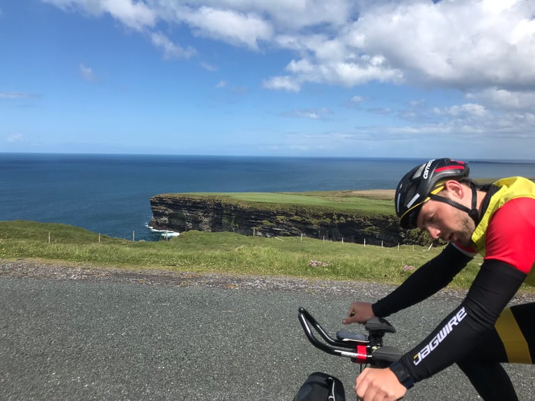 Rider tucked on a Brompton's aero bars passes a grassy sea cliff on Ireland's west coast during the TransAtlantic Way.