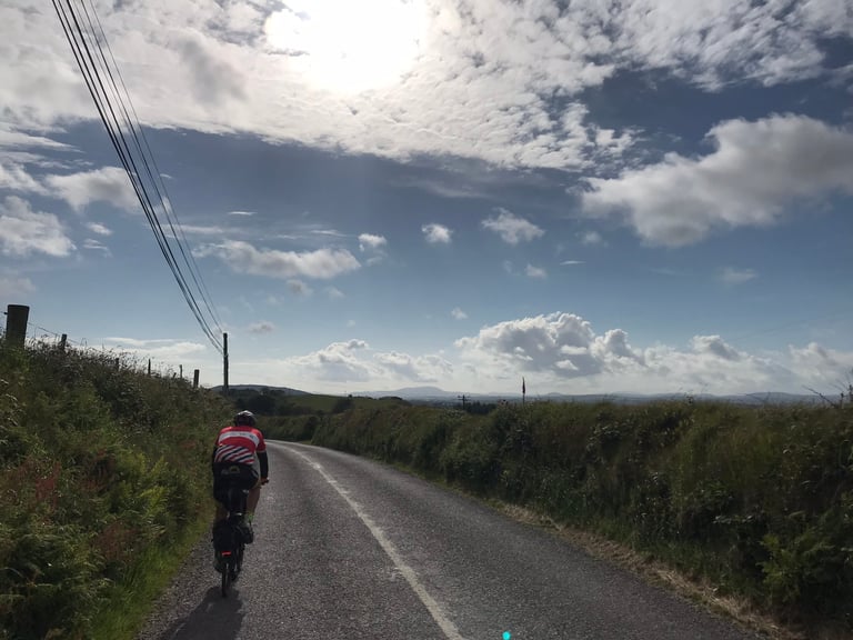 TransAtlantic Way rider on a loaded Brompton cycles a hedged Irish lane under blue sky near the end of the race.