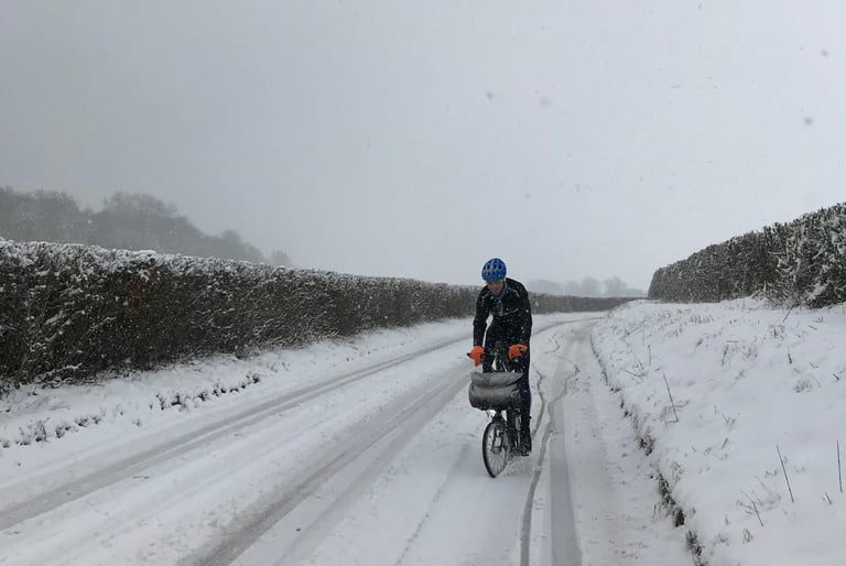 Rider on a Brompton with a bar bag rides a snow-covered lane between hedges in a training ride for the TransAtlantic Way.