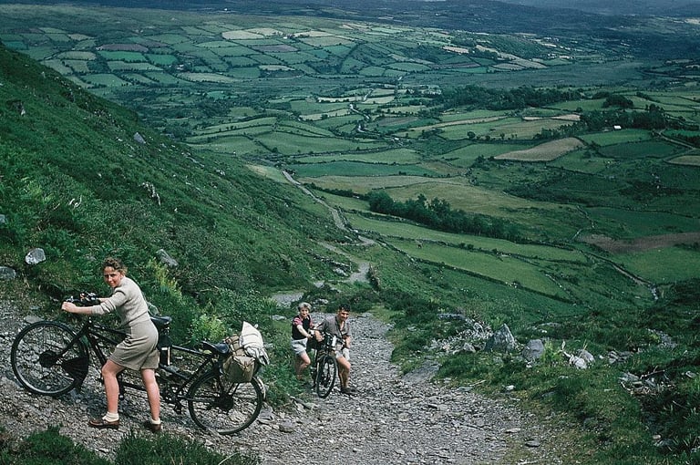 Early Rough Stuff Fellowship members push touring bikes up a steep stony track above patchwork fields in Southern Ireland.