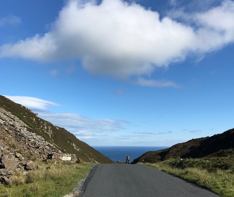 A lone rider at the crest of a quiet road through a rocky gap, the Atlantic beyond, on day two of the TransAtlantic Way.