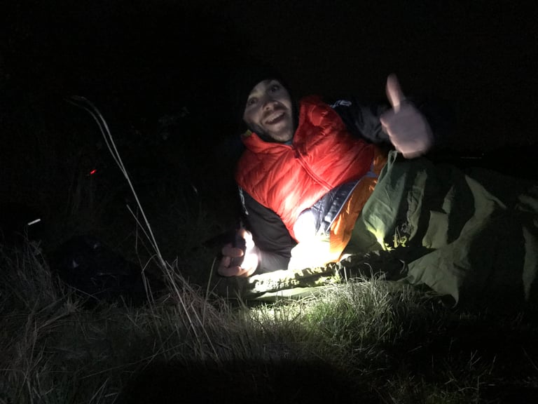 Rider in a red jacket grins and gives a thumbs up from a bivvy bag on grass at night during a bikepacking training trip.