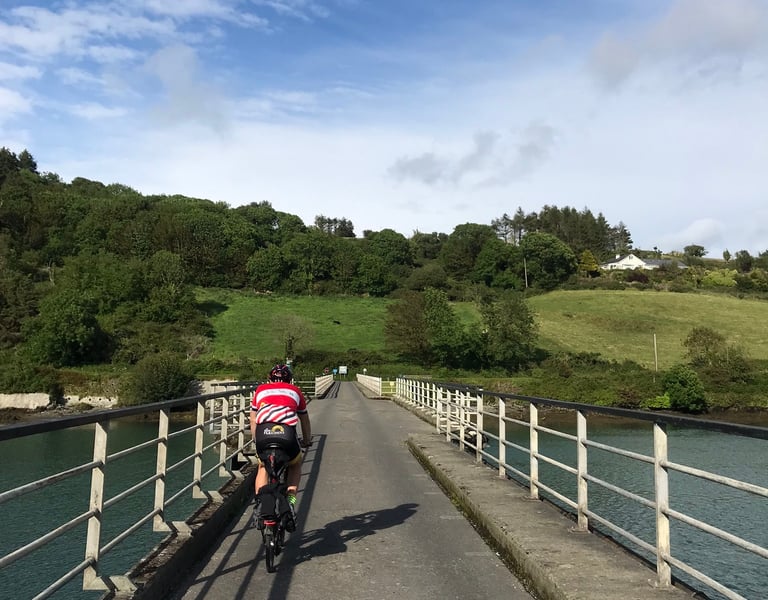 Brompton rider crosses a narrow bridge over an estuary towards green wooded hills on the TransAtlantic Way.