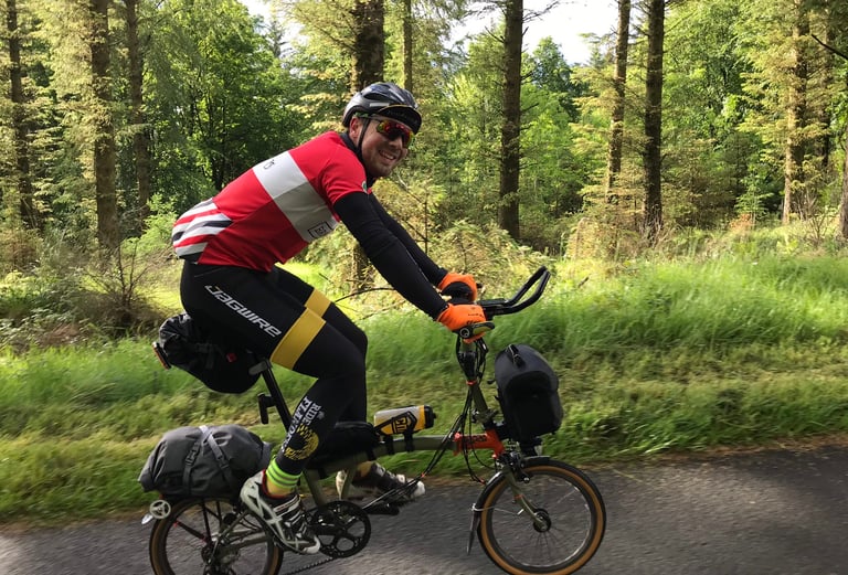 Smiling rider in a red jersey pedals a loaded Brompton along a forest road on day one of the TransAtlantic Way.