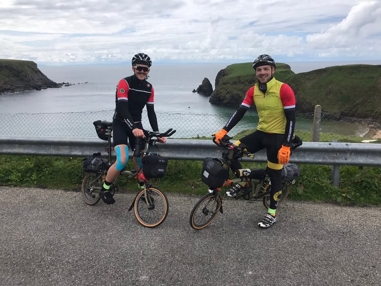 Mark and Jack pose with their loaded Bromptons beside a crash barrier above a sea inlet, one knee strapped with tape.