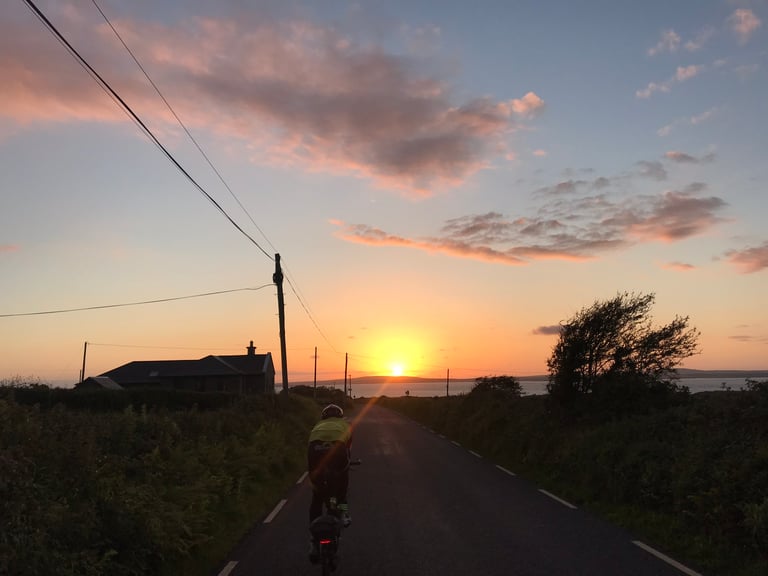 Brompton rider heads towards the setting sun on a narrow lane above the Atlantic, TransAtlantic Way Race.