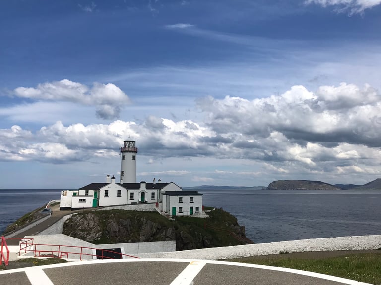 Fanad Head Lighthouse on a rocky headland in Donegal, with white keeper's buildings, sea and cloudy sky.